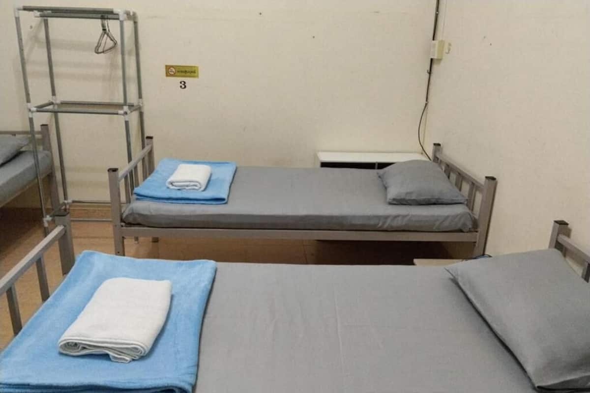 A dormitory room with two single beds, a metal shelf unit, and a wall-mounted electrical outlet.