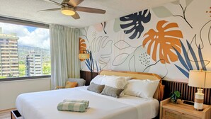 Une chambre d’hôtel avec un lit, un ventilateur de plafond, une table de chevet, une lampe, une fenêtre donnant sur des immeubles et un mur orné d’œuvres d’art abstraites.