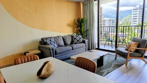 Un salon moderne avec un canapé, une table à manger et un balcon offrant une vue sur les bâtiments et les montagnes.
