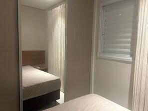 A bedroom with a bed, a bedside table, a window with blinds, and a door leading to another room. - Studio apartment in Tatuapé. 40 m2 (São paulo)