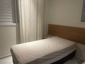 Uma cama de solteiro com cabeceira de madeira, mesa de cabeceira com abajur e janela com cortinas.