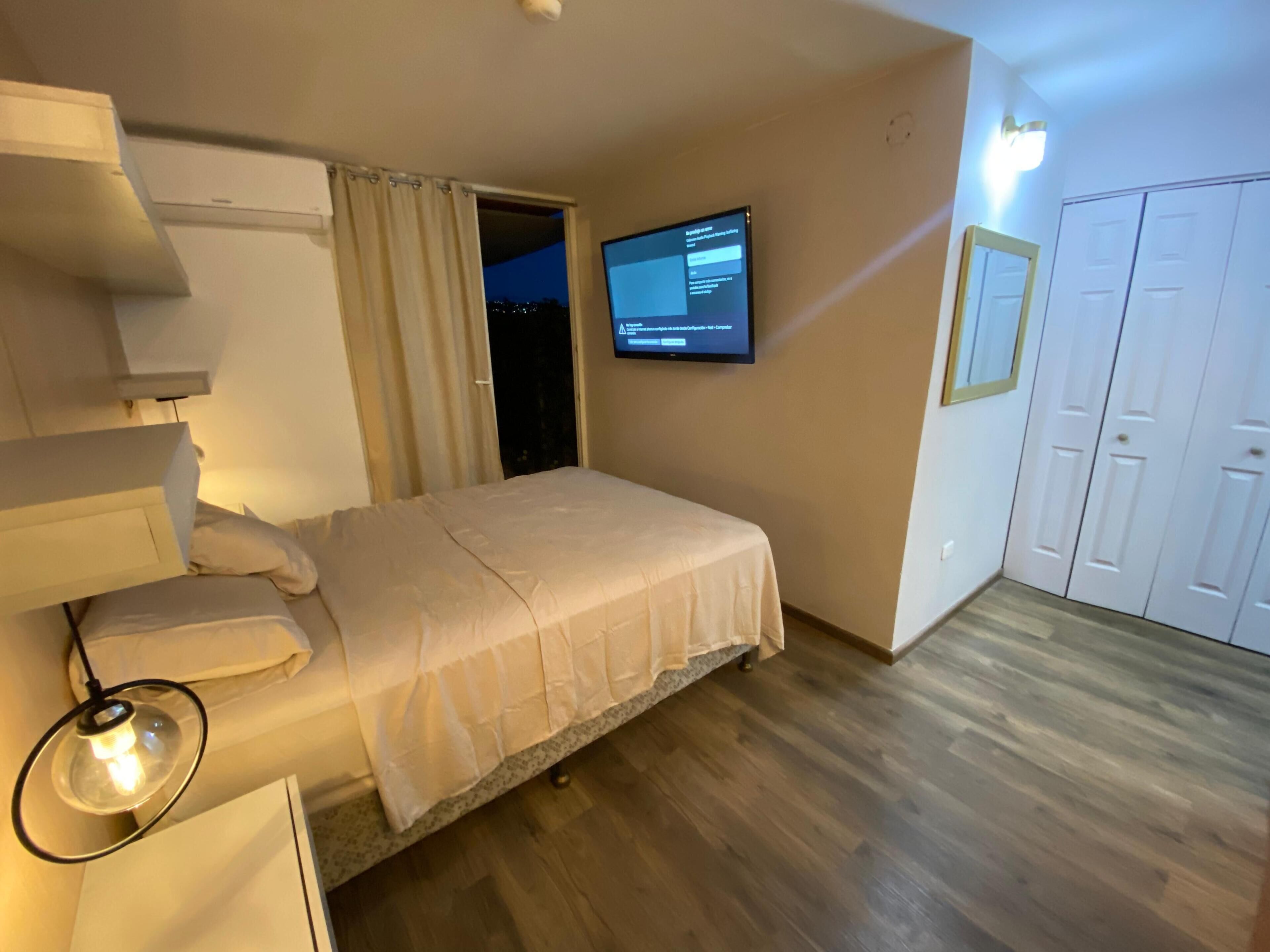 Un dormitorio con cama, televisor montado en la pared, armario y puerta.