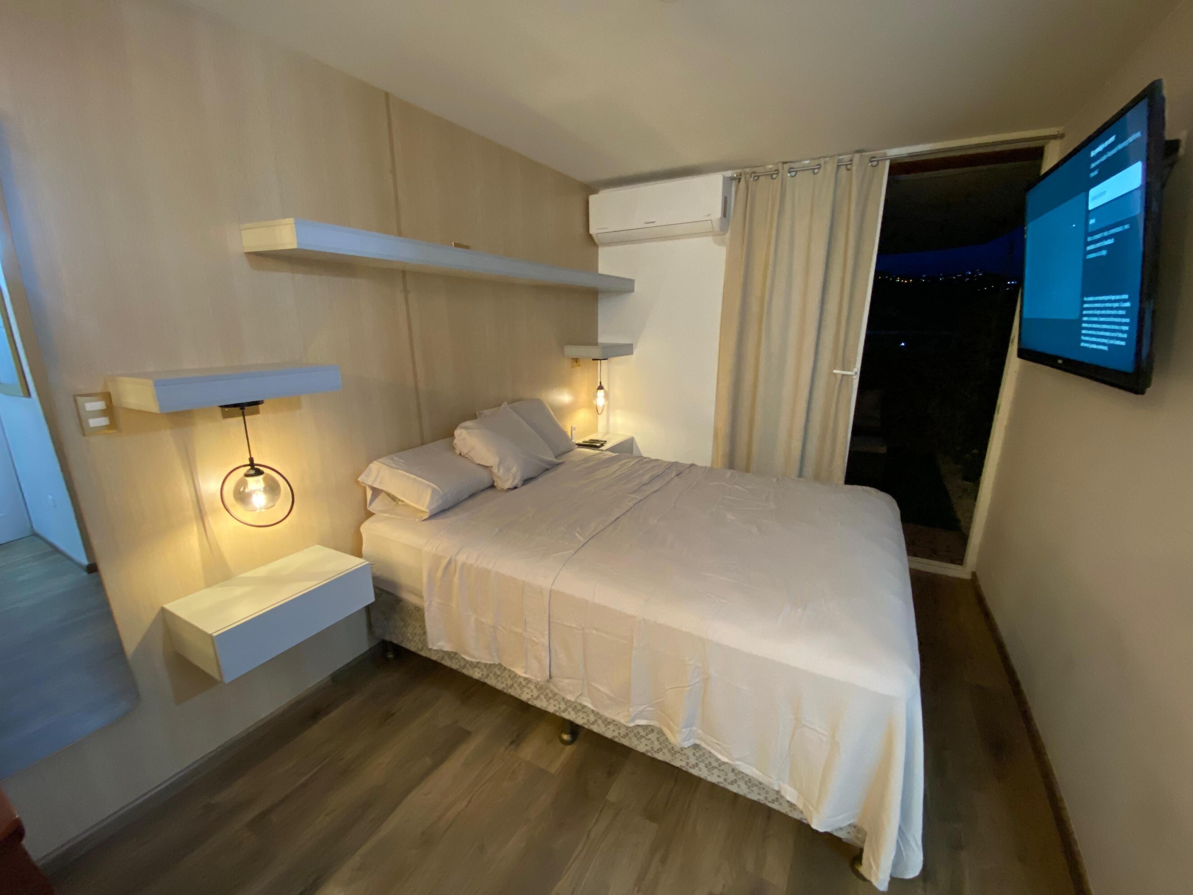 A modern bedroom with a bed, bedside tables, a mounted TV, and a view of the outside.
