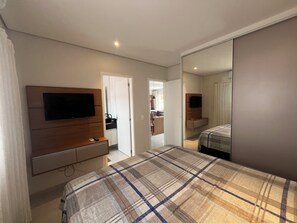 A bedroom with a bed, a TV on a wooden cabinet, a mirror, and a door leading to a bathroom. - Wonderful apartment 50 meters from the beach. (Bertioga)