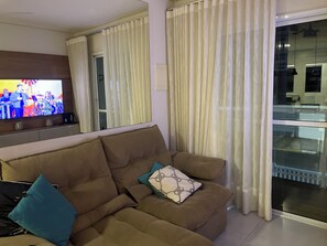 A living room with a brown sofa, teal and white cushions, a flat-screen TV, and sheer curtains. - Wonderful apartment 50 meters from the beach. (Bertioga)
