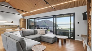 Ein modernes Wohnzimmer mit einer großen Eckcouch, einem kleinen Couchtisch und Blick auf Berge und Meer durch große Glastüren.