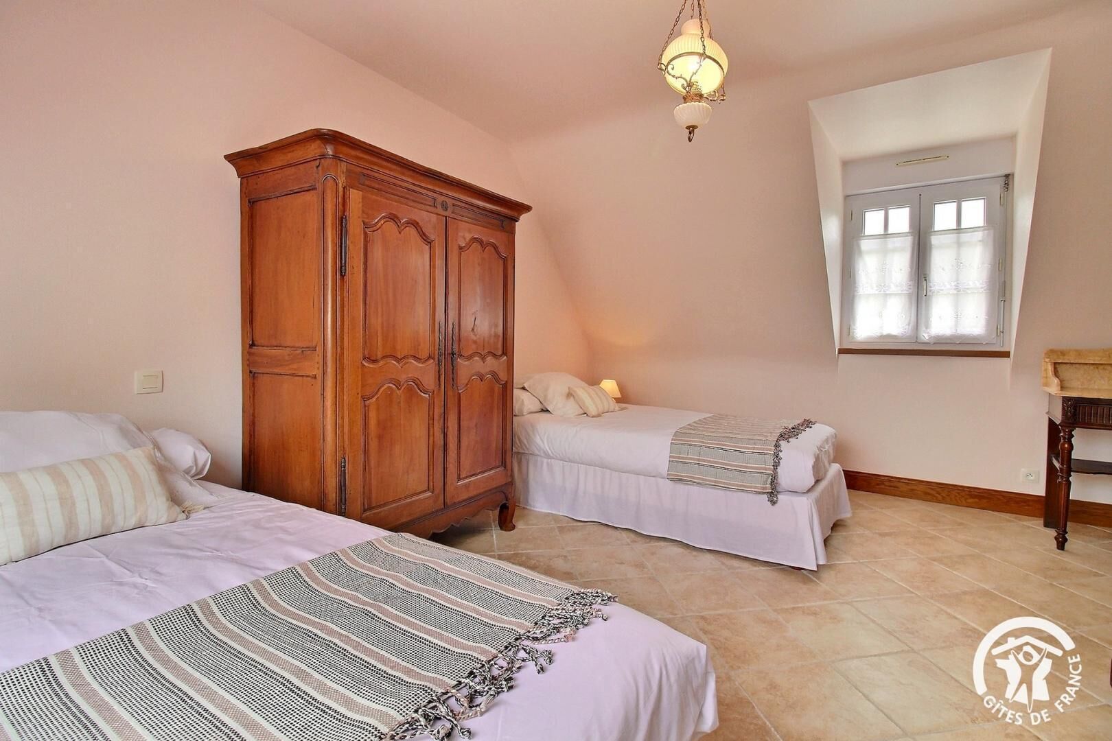 Ein Schlafzimmer mit zwei Betten, einem Holzschrank, einer Pendelleuchte und einem Fenster mit Vorhängen.