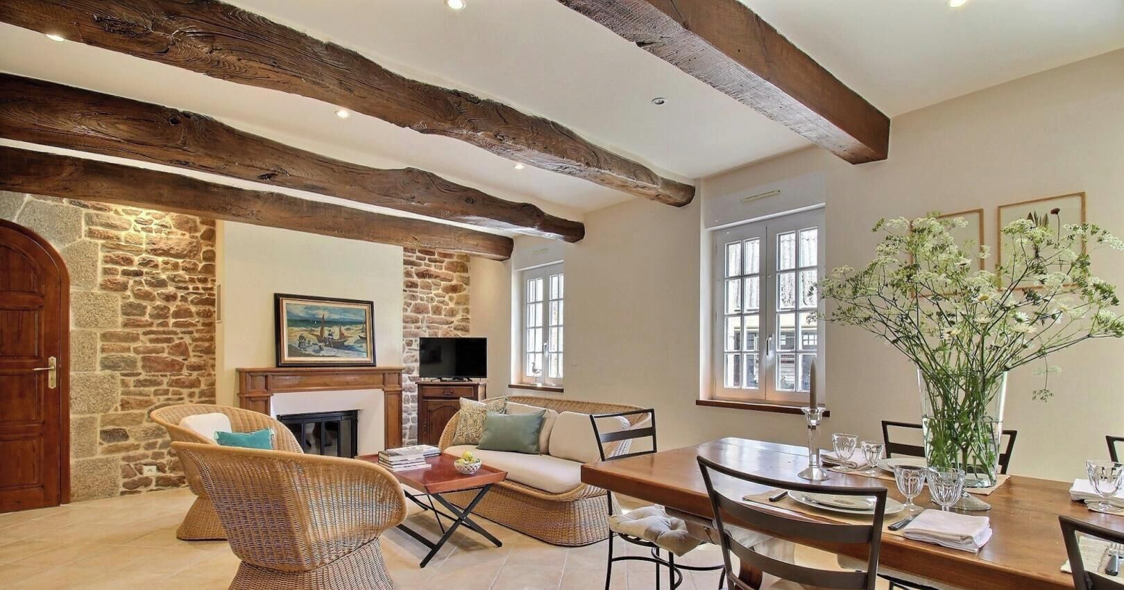 A cozy living room with a stone wall, wooden beams, wicker furniture, and a fireplace.