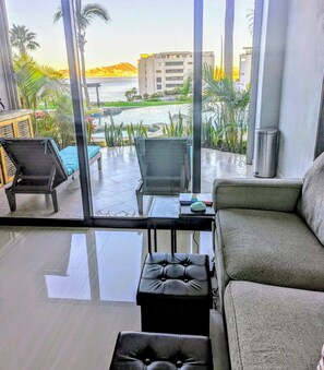 Ein modernes Zimmer mit Glastrennwand, einer Couch, einem Sessel, einem kleinen Tisch und Blick auf einen Pool und Gebäude.