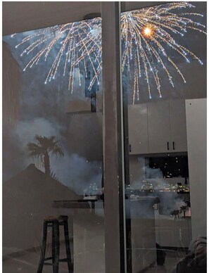 Ein Zimmer mit großem Fenster, einem Holzschemel und Blick auf Feuerwerk vor dem Fenster.