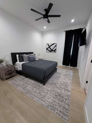 Ein modernes Schlafzimmer mit Bett, Nachttischen, Deckenventilator, Wand-TV und schwarzem Vorhang.