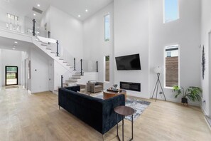 Ein geräumiger Wohnbereich mit einem schwarzen Sofa, einem Couchtisch und einem an der Wand montierten Fernseher. Eine Treppe führt zu einer oberen Ebene.