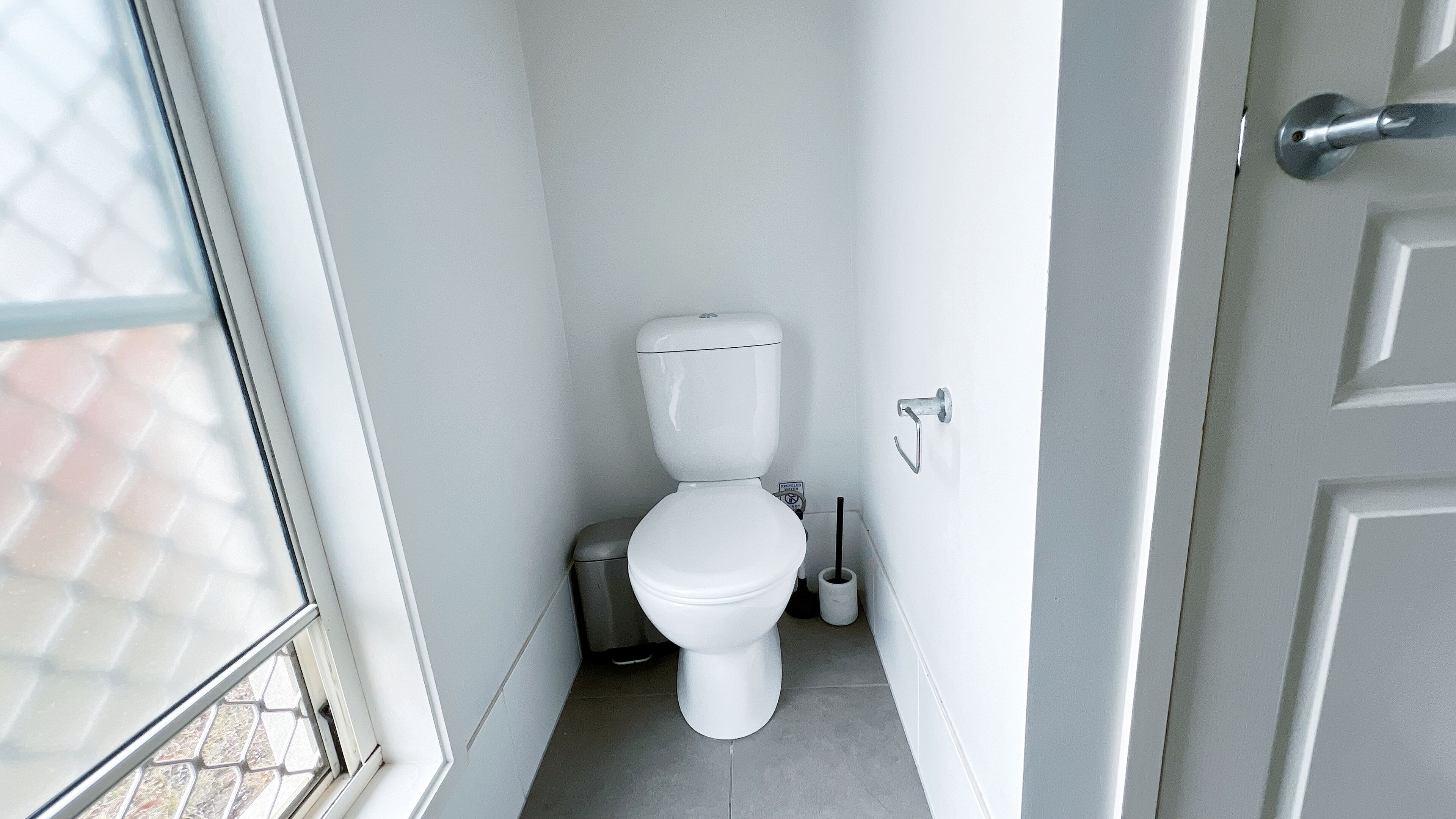 A small, narrow bathroom with a toilet, a trash bin, and a toilet brush.