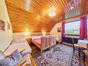 Une chambre mansardée chaleureuse, avec un plafond en pente, une grande fenêtre offrant une belle vue, un lit en bois, une petite table et un tapis à motifs.