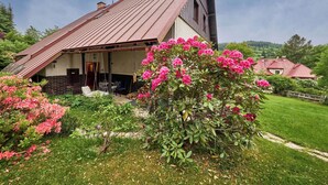 A house with a sloped roof, a porch with outdoor seating, and a garden with flowering plants. - Apartment Černý by Interhome (Rokytnice nad Jizerou)