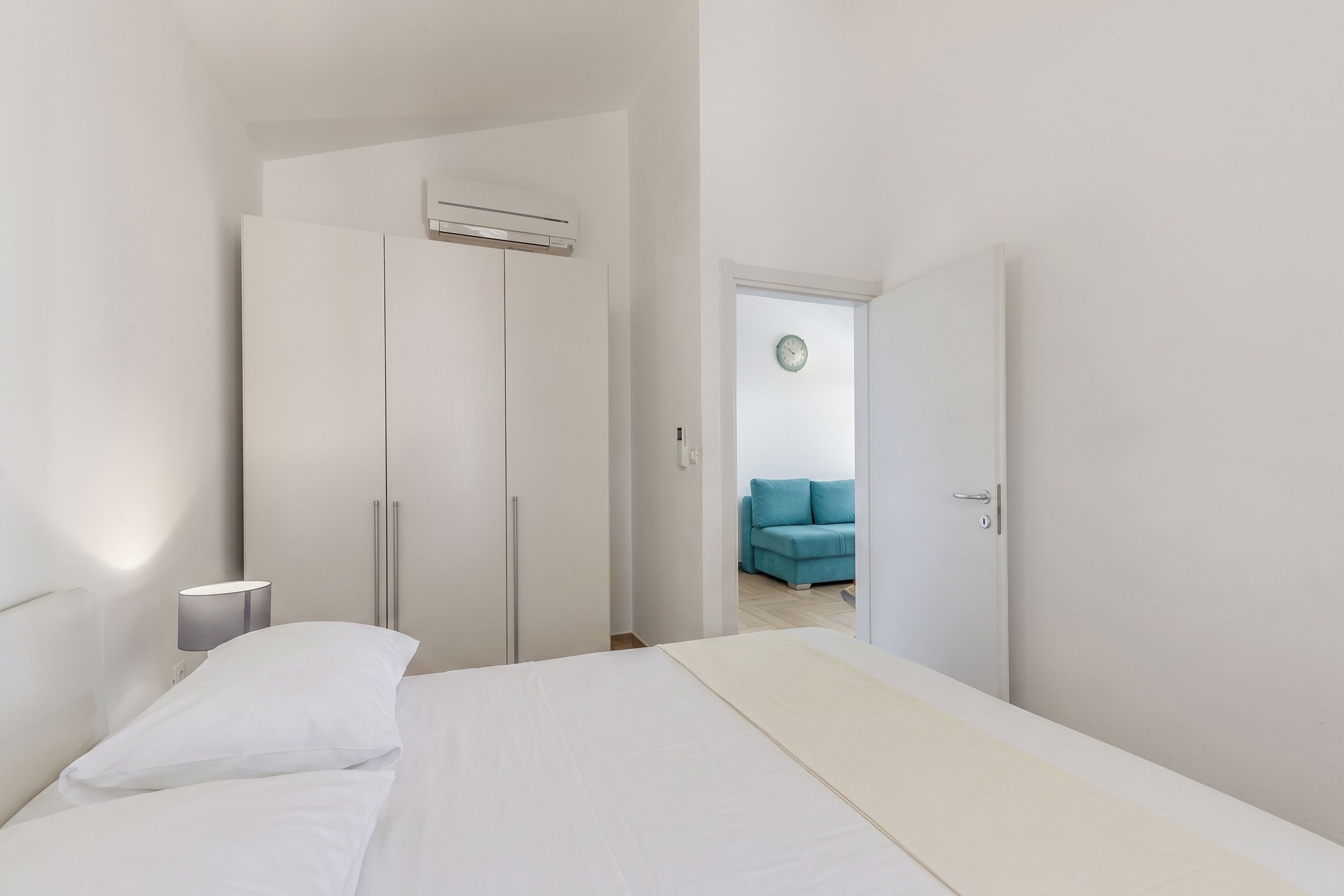 A bedroom with a bed, a wardrobe, a bedside lamp, and a view of a living area with a sofa.