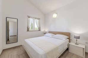 Ein Schlafzimmer mit einem Bett, einem Nachttisch, einem Wandspiegel, einem Fenster mit Vorhängen und einer Deckenleuchte.