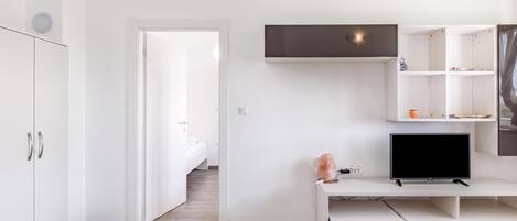 A modern, minimalist room with a bed, a desk with a TV, and a shelving unit.