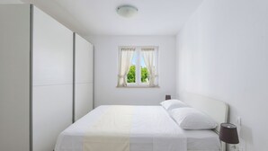 Een slaapkamer met een bed, nachtkastje, lamp, raam met gordijnen en een kledingkast.
