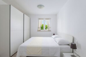 Ein Schlafzimmer mit Bett, Nachttisch, Lampe, Fenster mit Vorhängen und einem Kleiderschrank.
