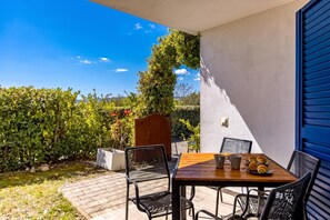 Une terrasse extérieure avec une table dressée pour deux personnes, entourée de chaises, et donnant sur un jardin agrémenté d’une structure en bois.