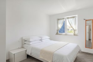 Ein Schlafzimmer mit einem Bett, einem Nachttisch, einem Fenster mit Vorhängen und einem Spiegel.