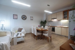 Ein modernes Wohnzimmer mit Esstisch, weißen Möbeln und Küchenzeile mit Holzausbau und integriertem Backofen.