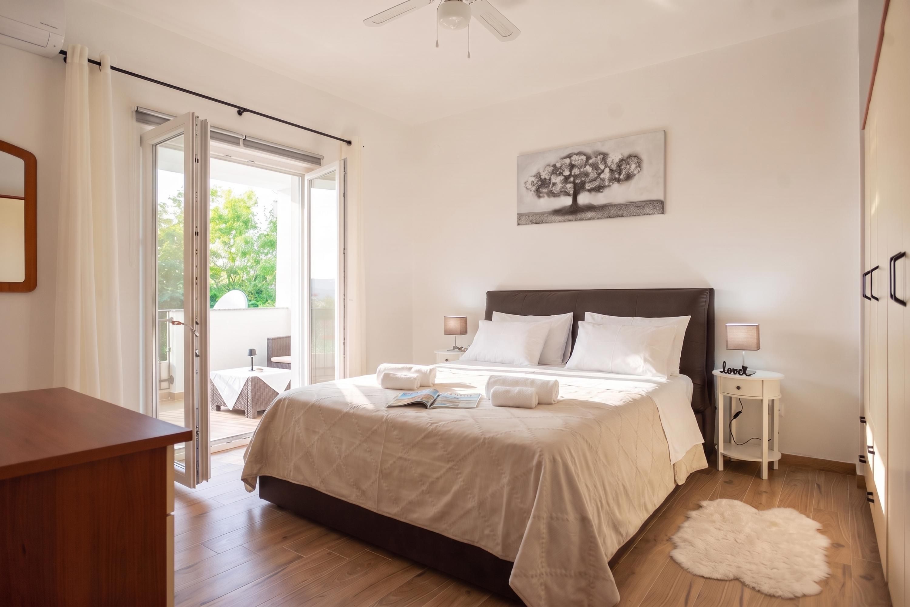 A bedroom with a bed, a bedside table, a lamp, a painting, and a view through a glass door.