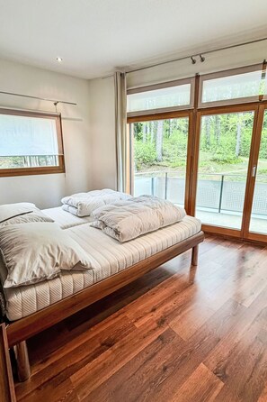 Ein modernes Schlafzimmer mit einem hölzernen Bett, großen Fenstern und Blick auf Grünflächen.