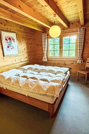 A wooden cabin bedroom with a large bed, a hanging light fixture, and a window with curtains. - Zum Hirsch - upper floor by Interhome (Bellwald)