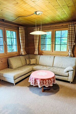 Um quarto aconchegante com sofá de canto, mesa redonda com toalha de mesa estampada e grandes janelas com cortinas.