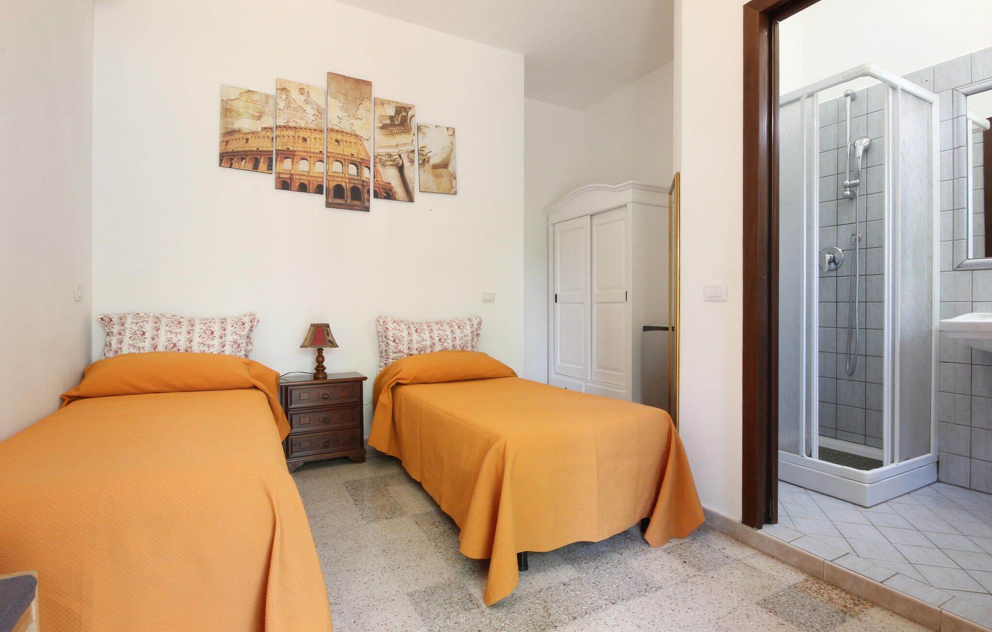 A bedroom with two beds, a nightstand, a lamp, a wardrobe, and a shower area.