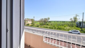 Ein Balkon mit Metallgeländer, der auf eine Straße mit einem Auto, ein Gebäude und freie Felder blickt.