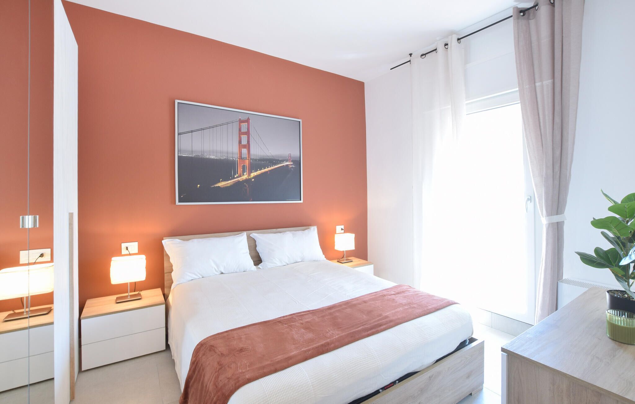 A hotel room with a bed, bedside tables, a lamp, a painting of the Golden Gate Bridge, and a window with curtains.