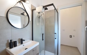 Ein Badezimmer mit rundem Spiegel, einem weißen Waschbecken, schwarzen Armaturen und einer Duschkabine aus Glas.