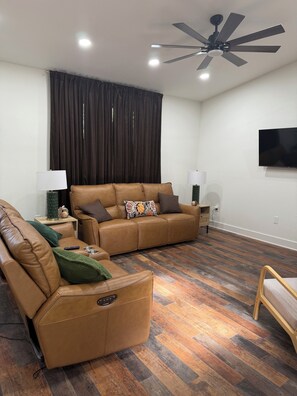 Ein modernes Wohnzimmer mit einem braunen Ledersofa, einem Holztisch, einer Stehlampe, einem Deckenventilator und einem an der Wand montierten Flachbildfernseher.