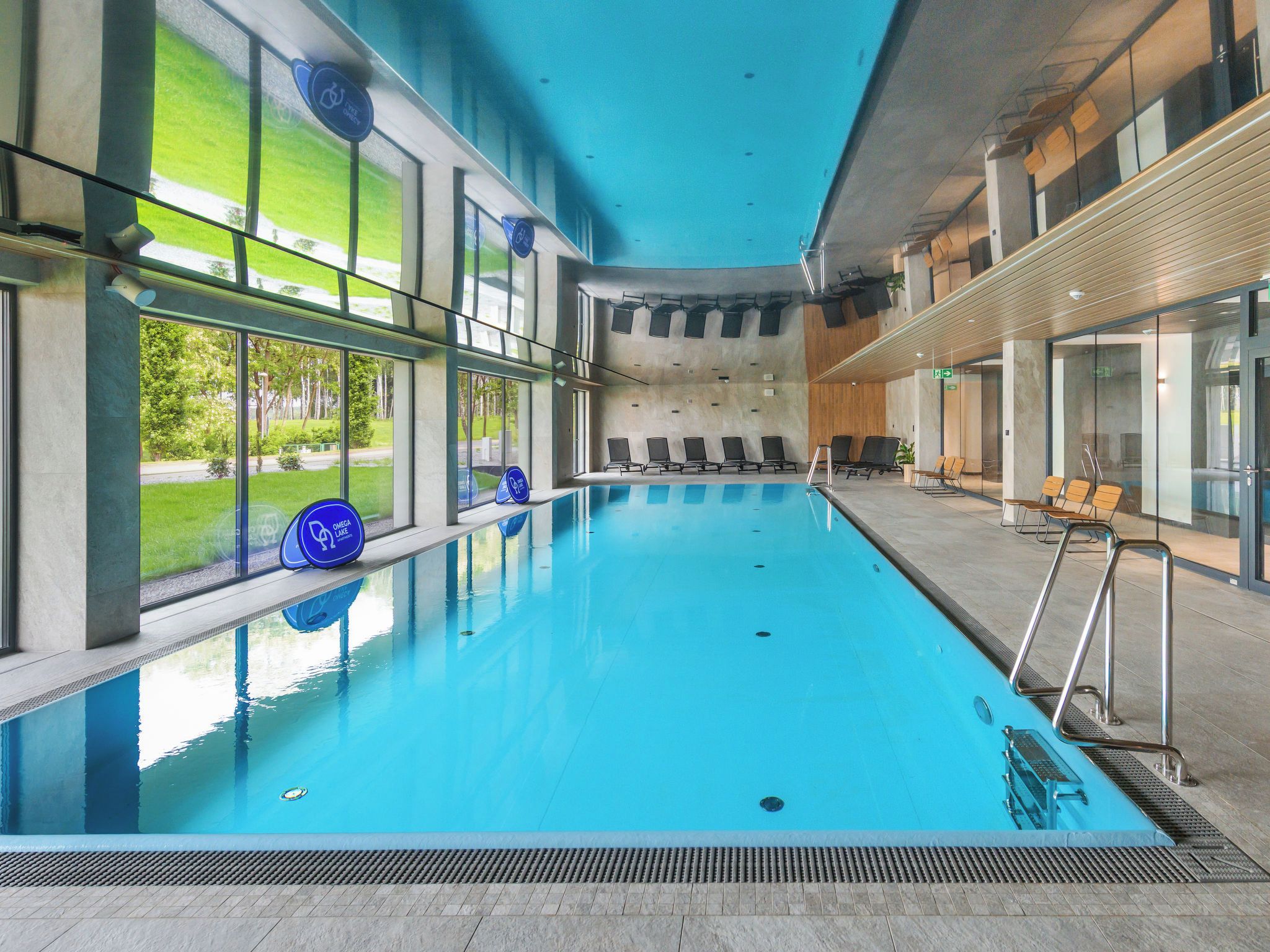 Une piscine intérieure avec des parois en verre transparent, de l’eau bleue et un espace détente avec des chaises.