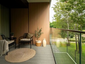 Een balkon met uitzicht op groen, een geweven vloerkleed, twee stoelen en een potplant.