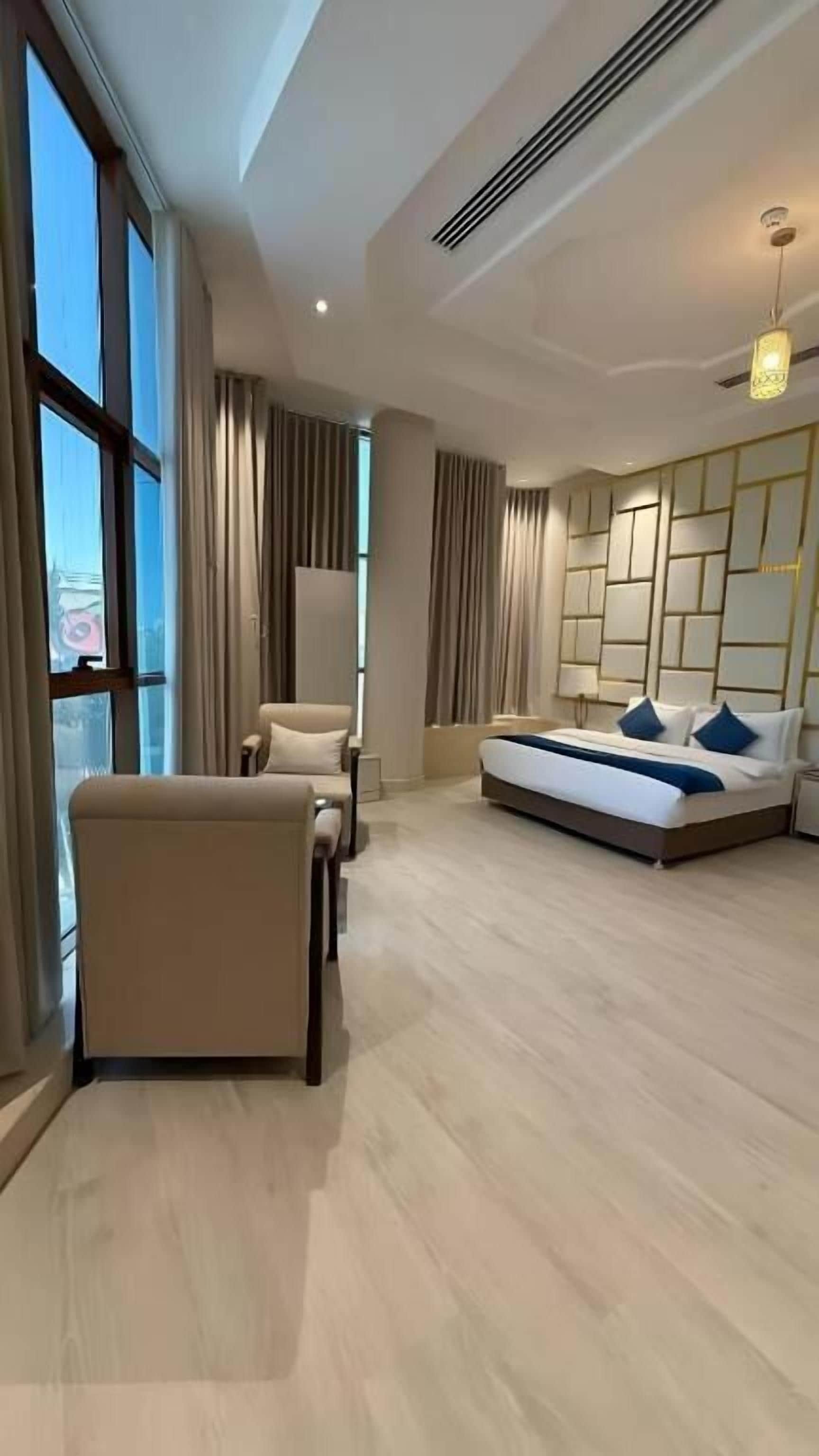 A modern hotel room with a large bed, a seating area with chairs, a desk, and a view through large windows.
