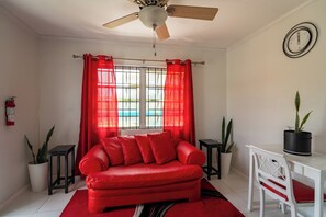 Ein Zimmer mit einem roten Sofa, einem Deckenventilator, einem Fenster mit roten Vorhängen, einem weißen Tisch und zwei Blumentöpfen.