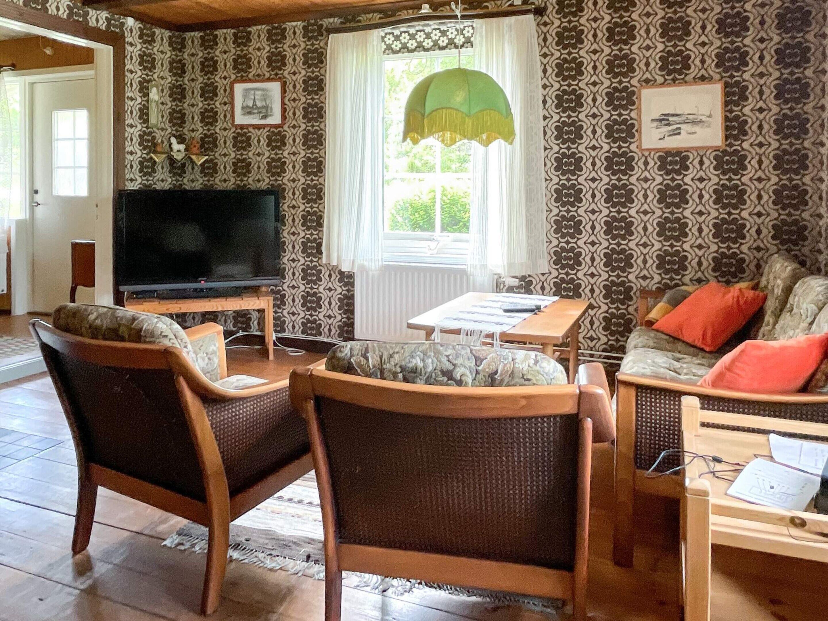 A living room with patterned wallpaper, a television, two wicker chairs, a small table, and a sofa with cushions.