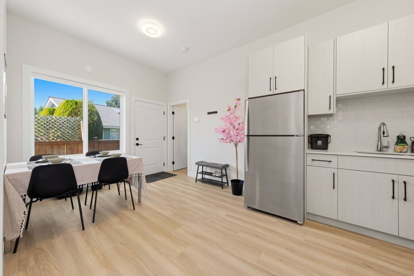 A modern kitchen with white cabinets, a stainless steel refrigerator, and a dining area with a table and chairs.