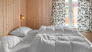 Una habitación con pared de madera, cama con ropa blanca y ventana con cortinas estampadas.
