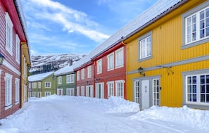 Eine schneebedeckte Straße gesäumt von bunten Häusern, ein klarer Himmel und ein Berg im Hintergrund.