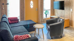 Una habitación con sofá, sillón, una mesita, televisor montado en la pared y una puerta con ventana.