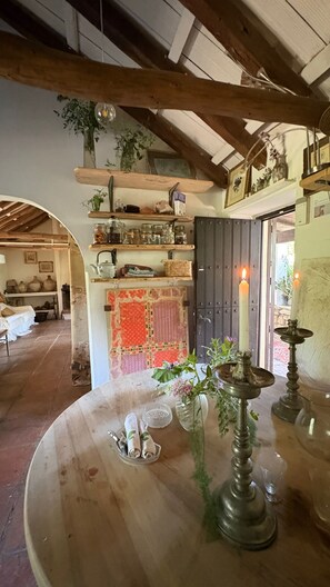 A rustic interior with exposed wooden beams, a wooden table with a vase of flowers, and shelves with various items.