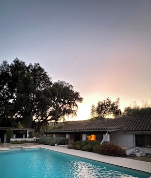 A swimming pool with a tiled deck, surrounded by greenery and a building with a tiled roof.