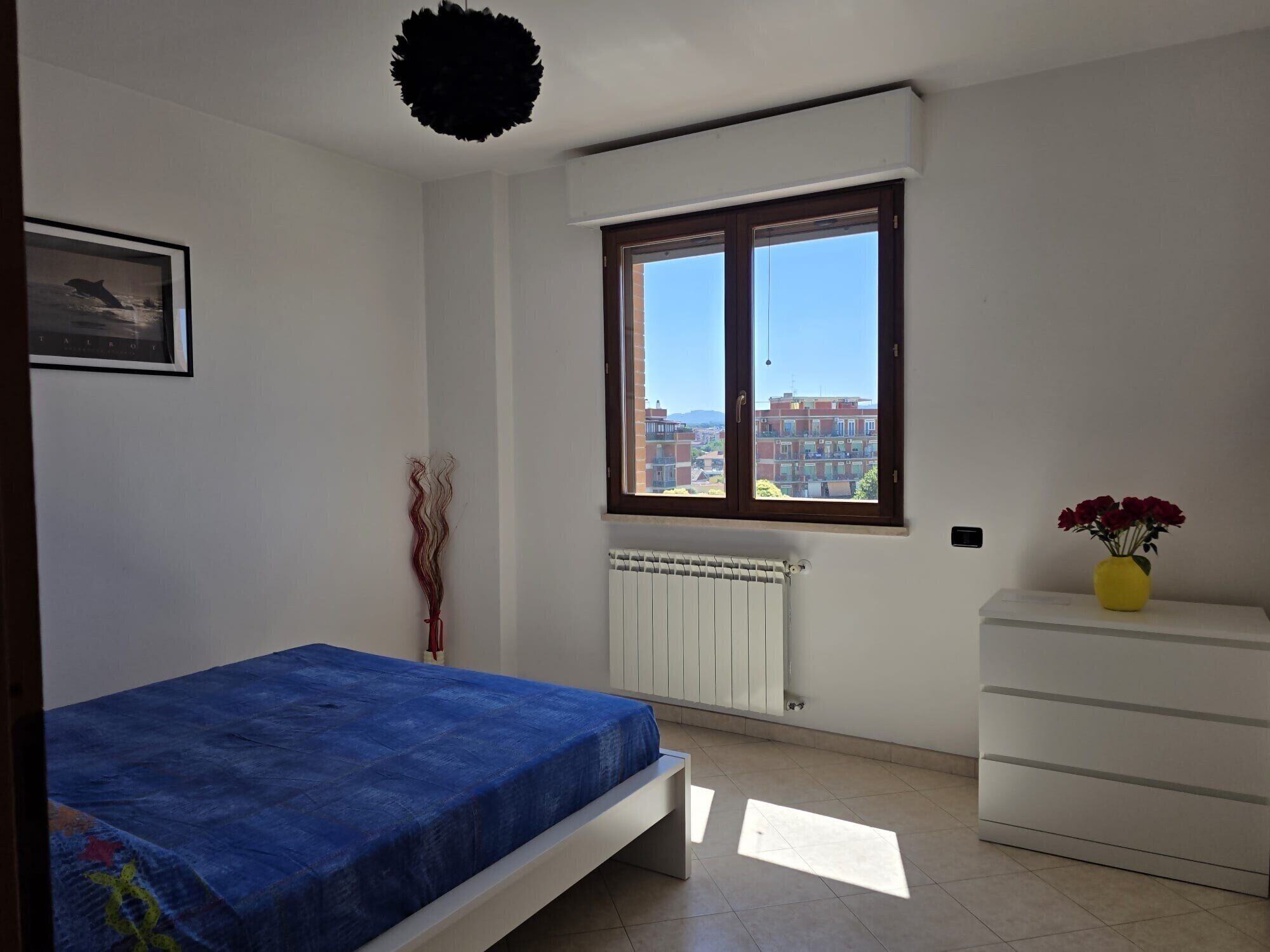 A bedroom with a bed, a radiator, a window with a view, a dresser, and a vase with flowers.