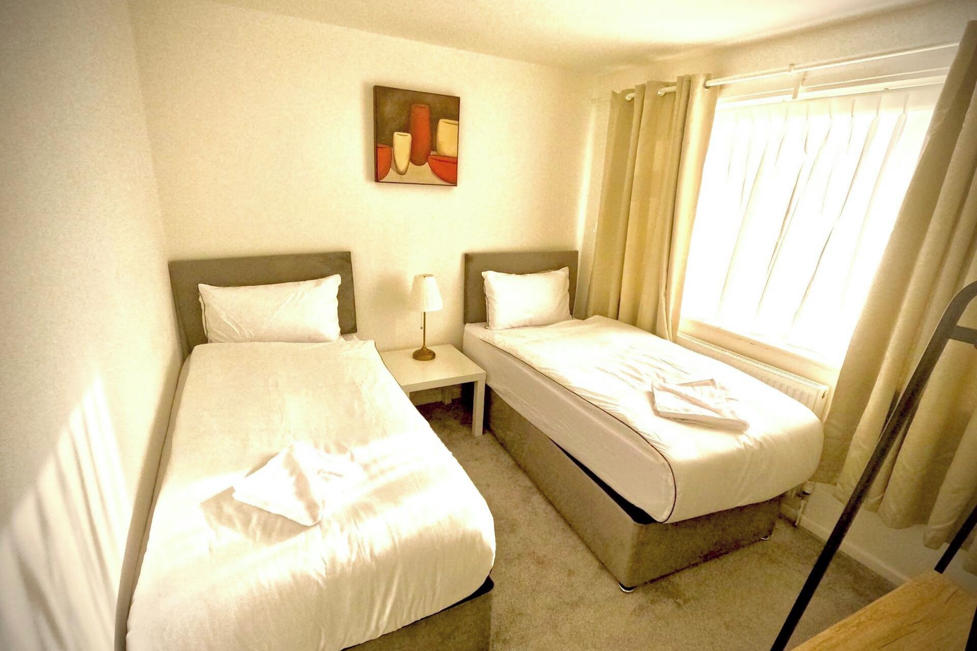 Een hotelkamer met twee enkele bedden, een nachtkastje met een lamp, een schilderij aan de muur en een raam met gordijnen.