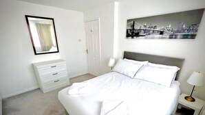 Habitación de hotel con cama, mesita de noche, lámpara, cómoda, espejo y una pintura de un paisaje urbano en la pared.